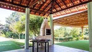 An outdoor patio with a barbecue grill and a table, surrounded by a covered area with a tiled floor and a wooden ceiling. - Chácara em Bragança Paulista com piscina e lazer (Bragança Paulista)