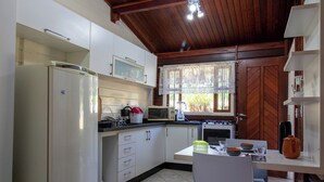A compact kitchen with white cabinets, a refrigerator, a microwave, and a stove. There is a window with lace curtains and a view of greenery outside. - Chácara em Bragança Paulista com piscina e lazer (Bragança Paulista)
