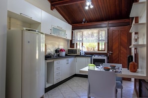 A compact kitchen with white cabinets, a refrigerator, a microwave, and a stove. There is a window with lace curtains and a view of greenery outside. - Chácara em Bragança Paulista com piscina e lazer (Bragança Paulista)