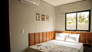 Un lit bien fait, avec une tête de lit en bois, une fenêtre donnant sur des palmiers et trois tableaux encadrés accrochés au mur.