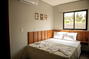 A neatly made bed with a wooden headboard, a window with a view of palm trees, and three framed pictures on the wall. - Apto com Piscina e Salão de Jogos a 100m da praia (Japaratinga)