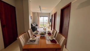 A dining area with a wooden table set for four, chairs, and a view of a balcony with a plant. - Apto Vista Mar a 100m da Praia de Japaratinga (Japaratinga)