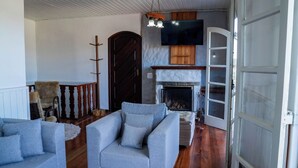 A cozy living room with a fireplace, a television mounted above it, and a large window. - Casa com lareira, Wi-Fi e varanda com vista (Campos do Jordão)