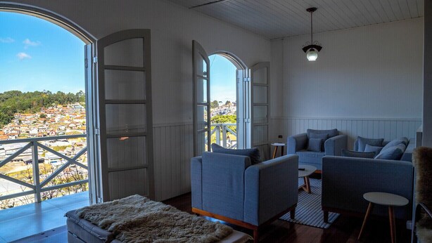 A spacious room with a large window offering a view of a coastal town. There are two sofas and a small table, with a fur throw on the floor. - Casa com lareira, Wi-Fi e varanda com vista (Campos do Jordão)
