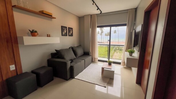 A modern living room with a sofa, ottomans, a small table, and a TV. There is a large window with a view of a beach and palm trees. - Apt Vista Mar 100m da Praia Piscina 2 Suítes (Japaratinga)