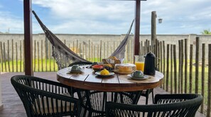 A round wooden table with plates, cups, and a jug of orange juice, surrounded by wicker chairs, set outdoors with a view of a wooden fence and greenery. - Dunas de Amaro Chalet: 3 Suites, Pool & Viewpoint (Santo Amaro)