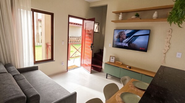 A living room with a sofa, a dining table with chairs, a TV mounted on the wall, and an open door leading to a balcony.