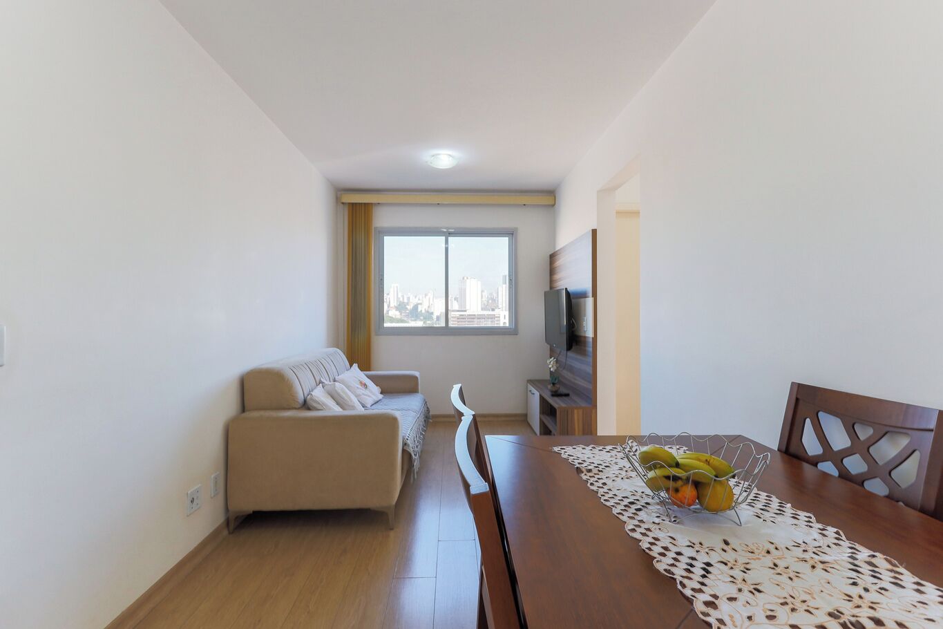 A modern living room with a sofa, a dining table with a fruit bowl, a TV, and a city view through the window.