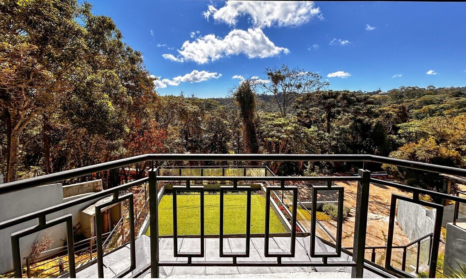 A view from a balcony with a metal railing, overlooking a grassy area and dense trees.