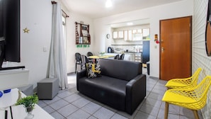 Een woonkamer met een zwarte bank, een televisie, een kleine tafel met een plant, en een keuken op de achtergrond.