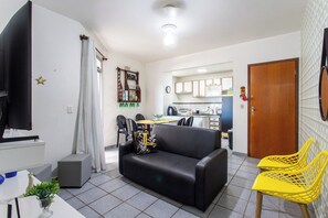 A living room with a black sofa, a TV, a small table with a plant, and a kitchen in the background. - Apto Pet Friendly com Garagem na Enseada Azul (Guarapari)