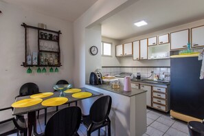 A compact kitchen with a dining area, featuring a small island, a clock, and basic appliances. - Apto Pet Friendly com Garagem na Enseada Azul (Guarapari)