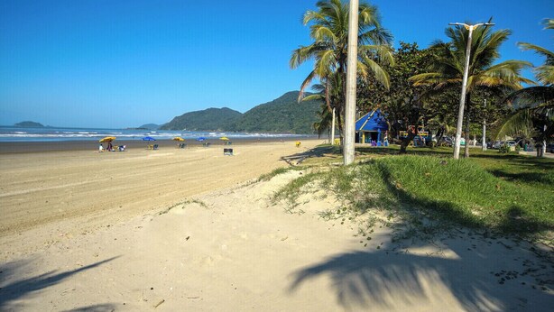 A beach with a clear sky, palm trees, and a small building with a blue roof. - Casa com Piscina e Garagem a 550m da Praia (Peruíbe)
