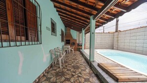 A covered outdoor area with a swimming pool, stone-tiled floor, and plastic chairs. - Casa com Piscina e Garagem a 550m da Praia (Peruíbe)