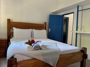 Ein ordentlich bezogenes Bett mit weißen Leintüchern und aufgerollten Handtüchern, ein hölzerner Kopfteil und eine blaue Tür im Hintergrund.
