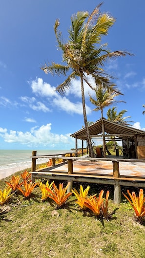 Ein Pavillon am Strand mit einer Holzterrasse, umgeben von tropischen Pflanzen und Palmen, mit Blick auf den Ozean.