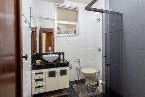 Uma casa de banho com um autoclismo branco, uma sanita preta com uma bacia branca, um espelho sobre a sanita e uma área de duche com porta de vidro.