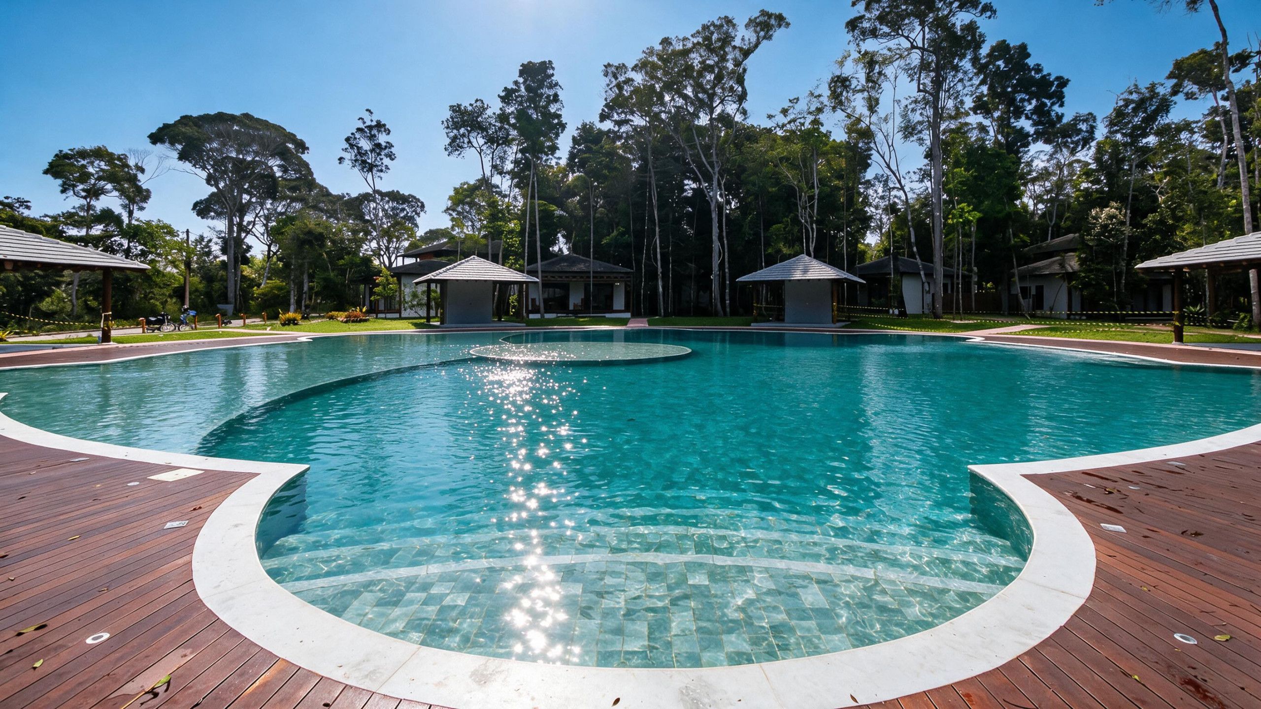 Una piscina blava i cristal·lina amb un pont de fusta al voltant, situada en un entorn tropical ple d'arbres i estructures cobertes.