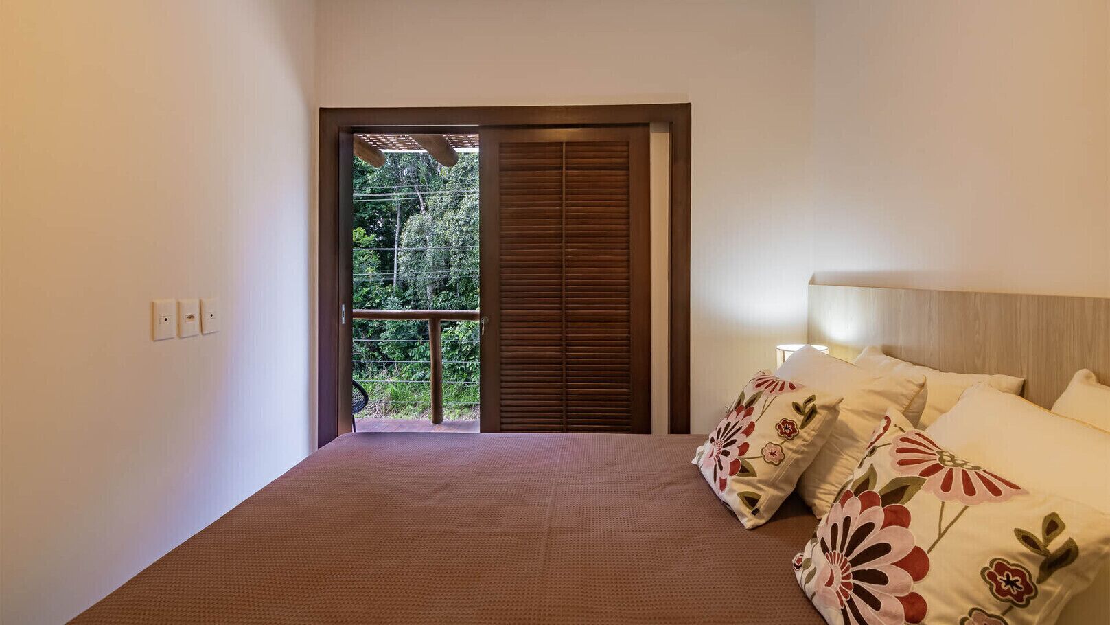 Una camera da letto con un letto, cuscini, un comodino e vista su un balcone con piante.