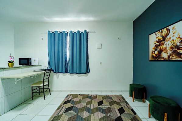 Une petite pièce propre, dotée d’une kitchenette, d’un coin repas avec une chaise et une table, d’un tapis à motifs, d’un rideau bleu et d’un tableau accroché au mur.