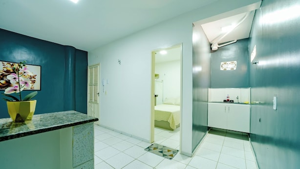 A hotel room with a bathroom, a bed, a small table with a vase of flowers, and a door leading to another room. - Flat com Wi-Fi a Poucos Metros do Aeroporto (Fortaleza)