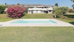 Ein Schwimmbad mit einer Rasenfläche davor, ein Gebäude mit einem Ziegeldach und ein Busch mit rosa Blüten auf der linken Seite.