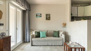 Ein Wohnzimmer mit einer Couch, einem Glastisch und einem Holznachttisch. Man sieht einen Heizkörper, eine Tür und ein Fenster mit Vorhängen.