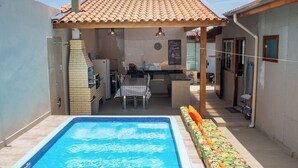 Un área junto a la piscina con techo de azulejos, una barbacoa, mesa de comedor y una pequeña cocina.
