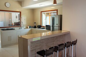 A modern kitchen with a central island, bar stools, a refrigerator, and a coffee maker. - Casa c Piscina e Churrasqueira na Praia Maranduba (Ubatuba)