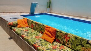 Una piscina con un sofá floral y cojines naranja, una fuente de agua y una planta en maceta.