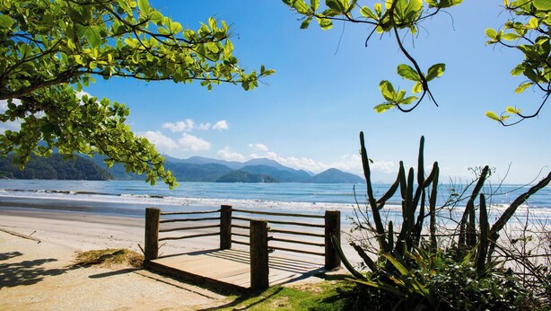 Une promenade en bord de mer, bordée d’une clôture en bois, qui mène à une plage d’où l’on peut admirer la mer et les montagnes au loin.