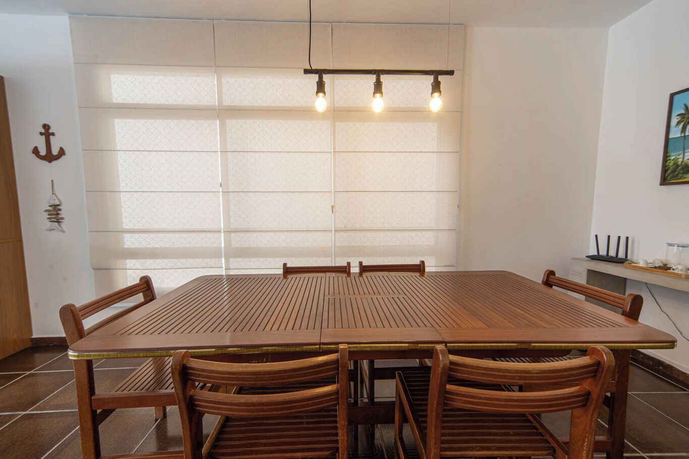 Ein Essbereich mit einem großen Holztisch und Stühlen, einer Lampe darüber und Blick ins Freie durch ein großes Fenster.