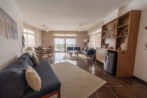 Ein modernes Wohnzimmer mit einem dunklen Sofa, einem Couchtisch und einem Essbereich mit Stühlen und Tisch. Es gibt eine Küchenzeile mit Holzausstattung und einem Kühlschrank.