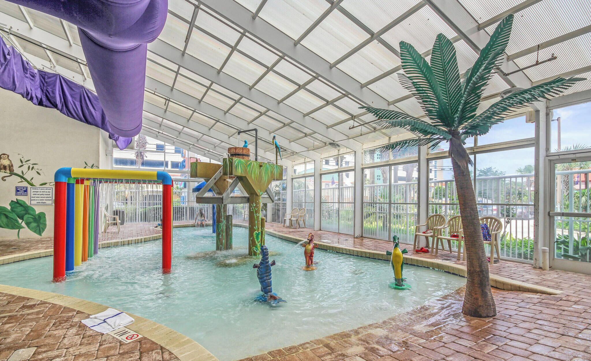 Un área de piscina interior con tobogán de agua, una palmera y una estructura de juegos para niños.