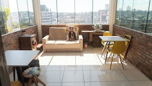 Ein modernes Wohnzimmer mit einer Couch, einem kleinen Tisch und gelben Stühlen. Große Fenster bieten Blick auf die Stadt.