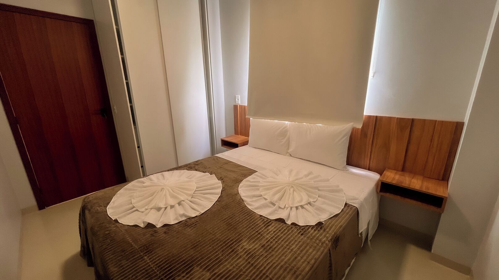 A hotel room with a bed, two decorative pillows, a wooden headboard, a nightstand, and a wooden door.