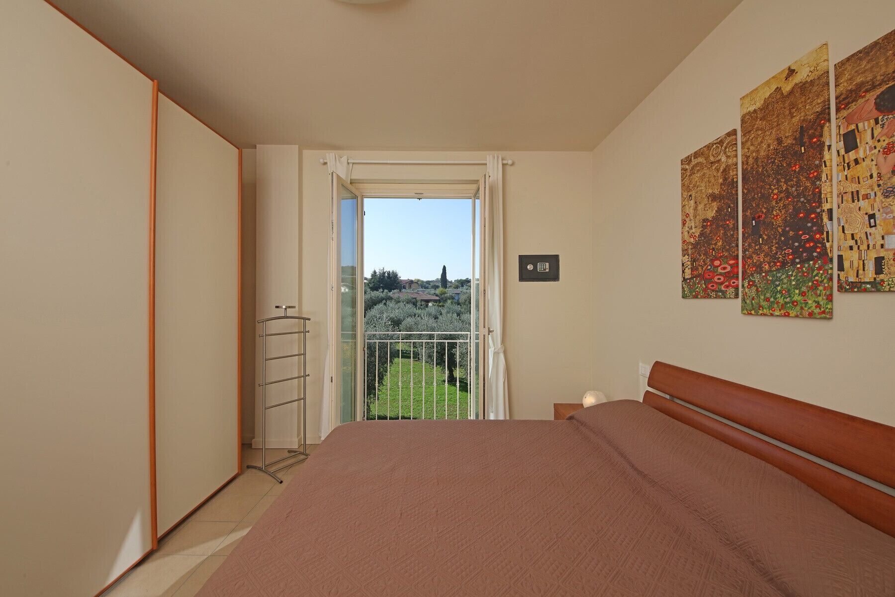 Een slaapkamer met een bed, een kledingkast, een balkon met uitzicht over de stad en twee schilderijen aan de muur.