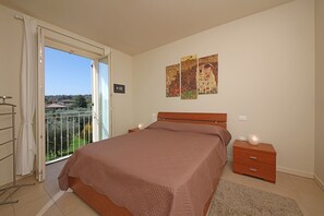 Ein Schlafzimmer mit einem großen Bett, einem Nachttisch, einer Wandleuchte und einem Balkon mit Blick auf Häuser und Grünflächen.