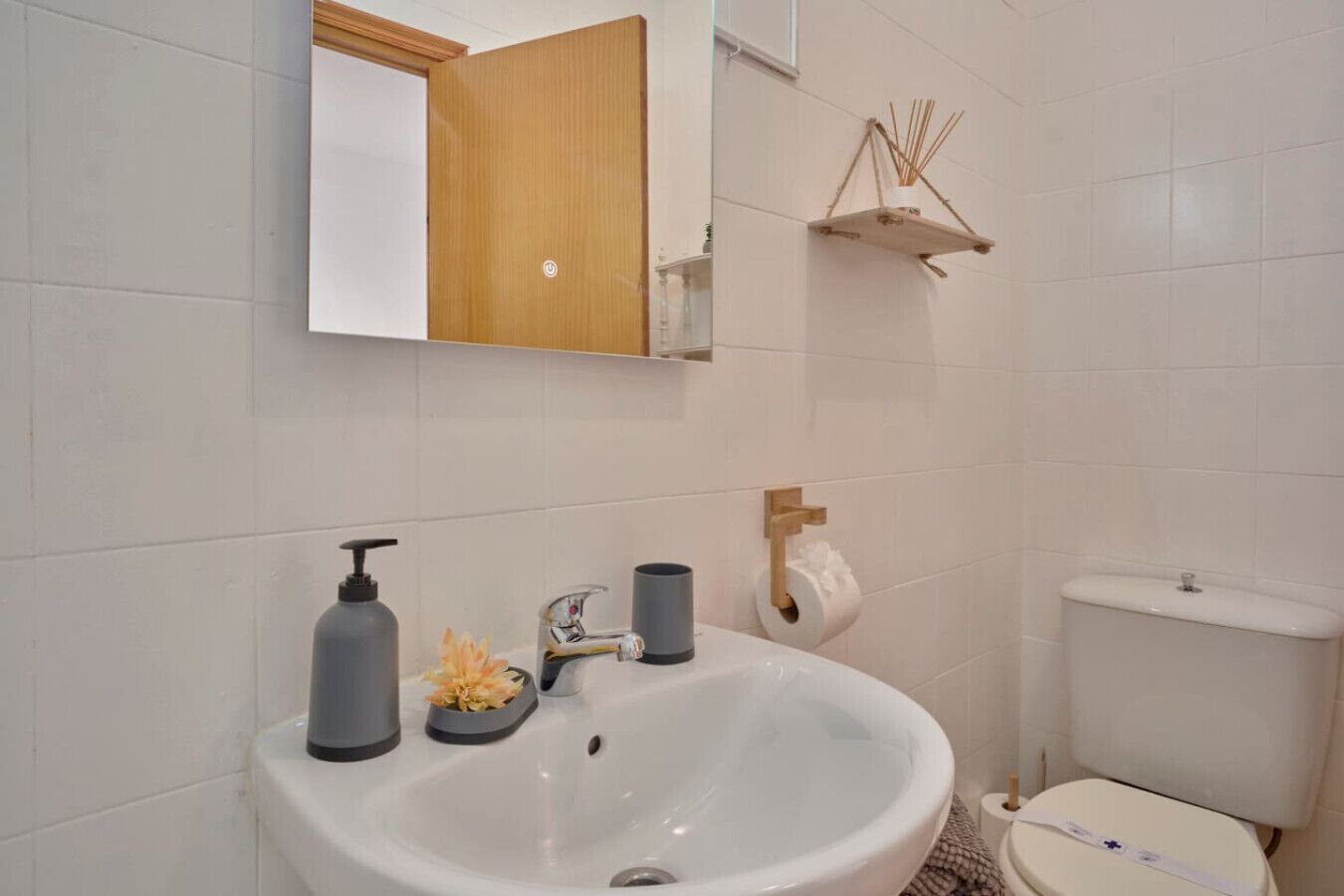 Une salle de bain avec un lavabo blanc, un miroir, des toilettes et une petite étagère sur laquelle se trouve un rouleau de papier toilette.