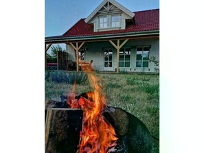 Een vuurplaats met vlammen, een houten terras en een huis met een rood dak op de achtergrond.