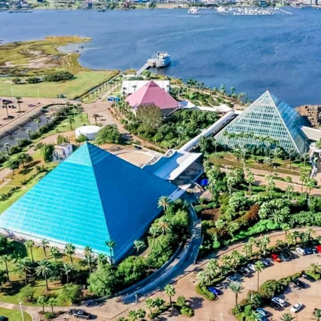 A large pyramid-shaped building with a blue roof, surrounded by greenery and a parking area.