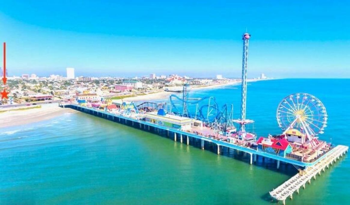 Vista aerea di un molo con ruota panoramica e giostre, che si protende su una distesa d'acqua con una spiaggia e uno skyline sullo sfondo.