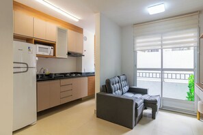 A modern kitchen with a built-in microwave, stove, and cabinets. There is a grey sofa and a small table in the living area, and a sliding glass door leading to a balcony.