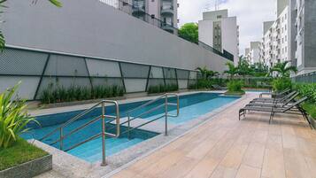Piscina com escadas e espreguiçadeiras, cercada por uma grade e vegetação.