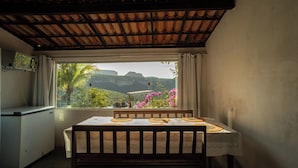 Een kamer met een tafel en stoelen, een raam met uitzicht op groen en bergen, en een betegeld plafond.