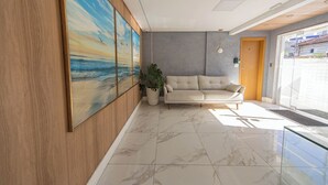 A modern interior with a large painting of a beach scene, a beige sofa, and a potted plant. - Apto com Ar e 2 Dorm na Vila Mirim perto da Praia (Santos)