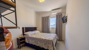 A bedroom with a bed, a nightstand, a TV mounted on the wall, and a window with curtains. - Apto com Ar e 2 Dorm na Vila Mirim perto da Praia (Santos)