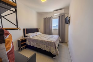 Ein Schlafzimmer mit einem Bett, einem Nachttisch, einem an der Wand montierten Fernseher und einem Fenster mit Vorhängen.