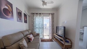 A living room with a sofa, a television on a wooden stand, and a view of the outside through a glass door. - Apto com Ar e 2 Dorm na Vila Mirim perto da Praia (Santos)