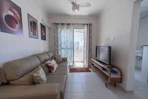 Ein Wohnzimmer mit einer Couch, einem Fernseher auf einem Holzständer und Blick ins Freie durch eine Glastür.